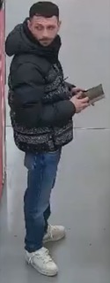 Man in a black puffer jacket and blue jeans reads a book.