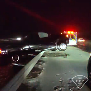 Damaged black car on a road, debris scattered, emergency lights visible