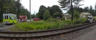 Ambulance and fire truck at accident scene. Car damaged, facing a house. Railroad tracks in foreground. Green foliage and trees.