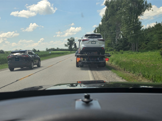 Car towing an SUV on a two-lane road, green field and trees in the background