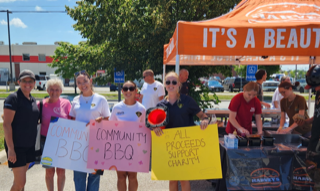 Group of people at a barbecue event. Signs read: COMMUNITY BBQ BBQ and ALL PROCEEDS SUPPORT CHARITY.