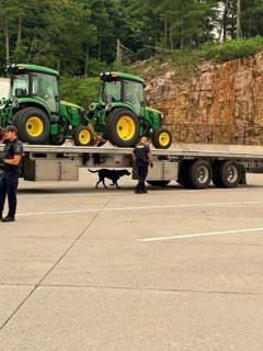Two John Deere tractors on a flatbed trailer, two people in uniforms, a black and white dog